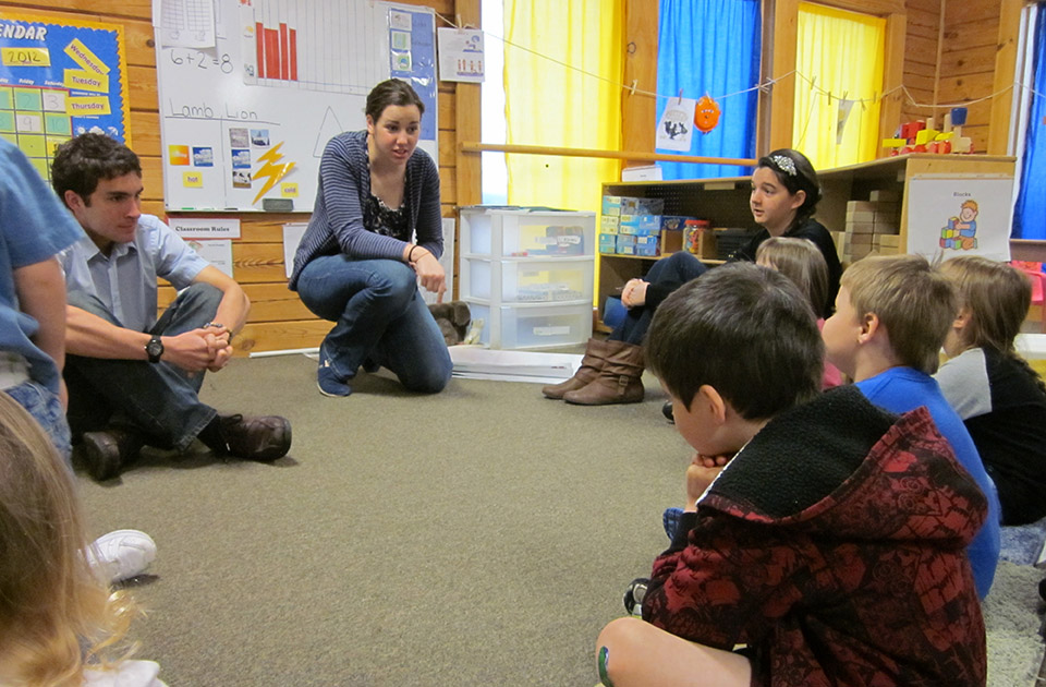 Students work at a local school. Students work at local school. Two SLU students sit on the floor at the front of a classroom with younger students gathered in a semi-circle.