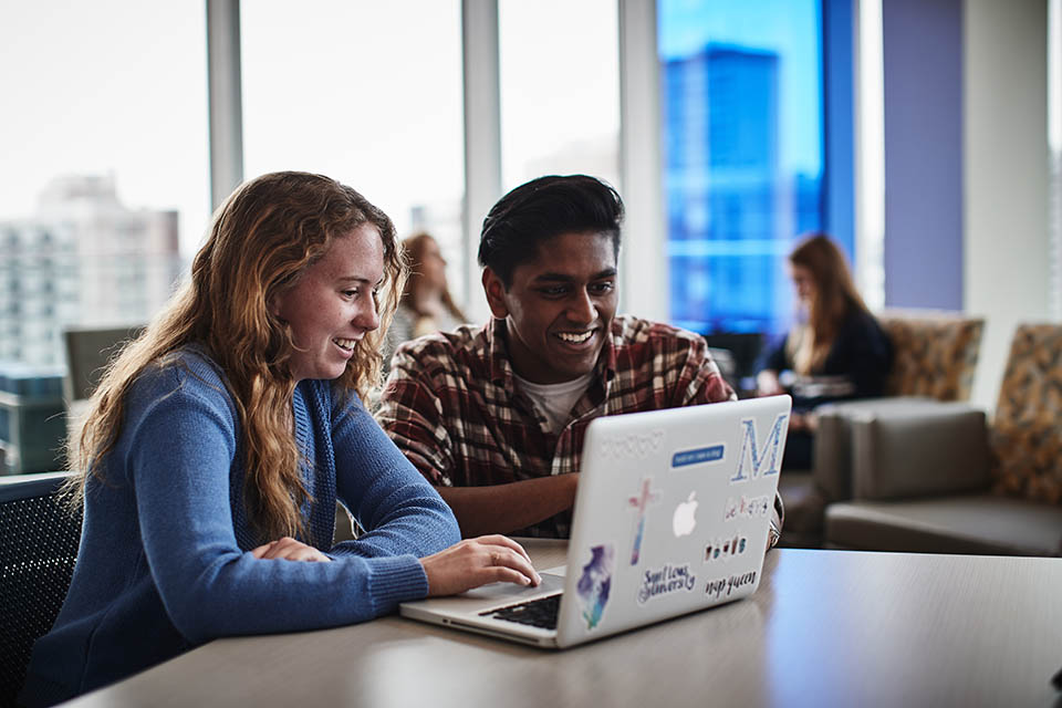 A female and male student sit at a computer in large room of other students, studying.