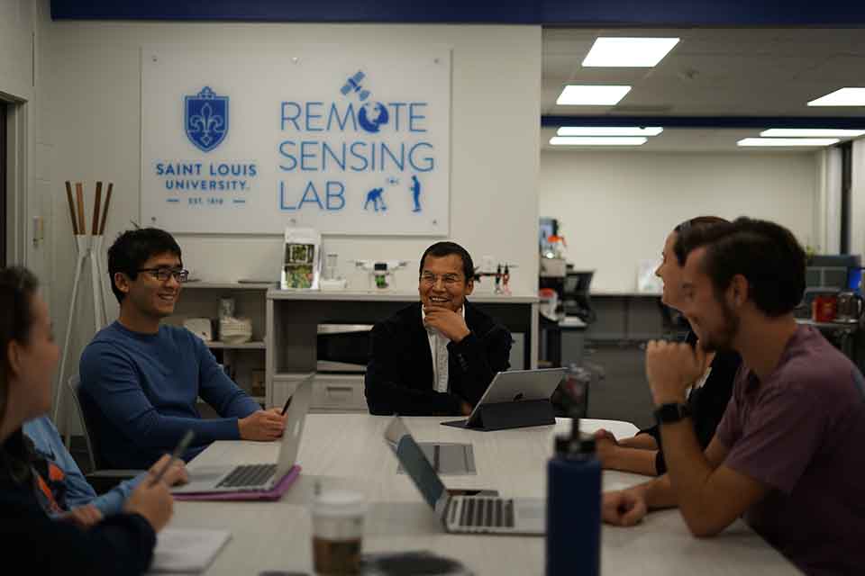 Five people sit around a table while working on their laptops. A white and blue sign in the background reads Saint Louis University Remote Sensing Lab.