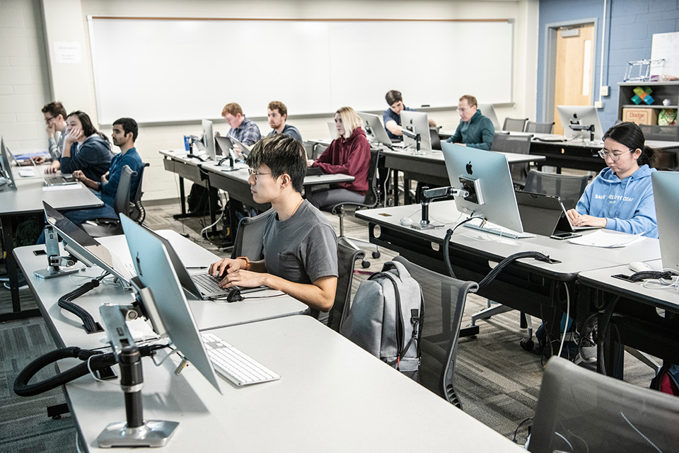 A computer science class taught in Ritter Hall. Students sit at desks in a classroom with laptops and computer monitors.