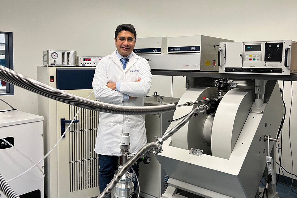 Reza Dastvan, Ph.D., in his lab. Reza Dastvan, Ph.D., in his lab, wearing a lab coat and standing next to some large pieces of equipment.