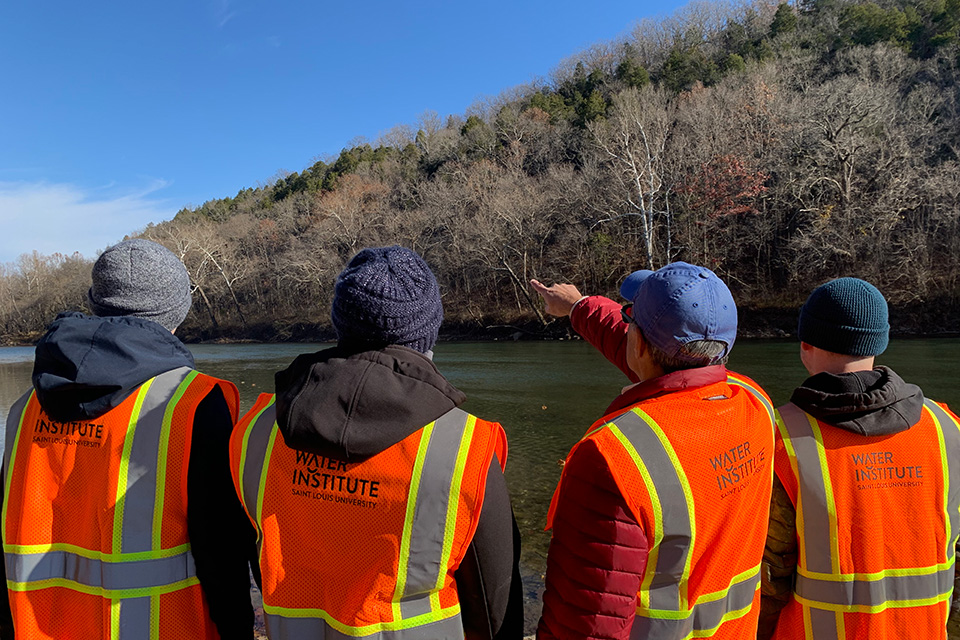 The WATER Institute team surveys the area before beginning their fieldwork. Four team members are shown from behind, wearing reflective vests that read Water Institute. One person points toward the distance as they look across the river at a shoreline of leafless trees.