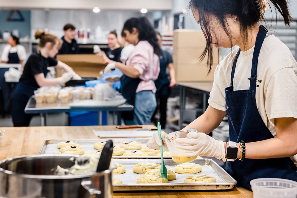 A student at work in Fresh Gatherings Cafe + Kitchen. A student wearing an apron brushes melted butter from a measuring cup onto pastries on a pan while standing at a kitchen counter. Other students are at work in the background.