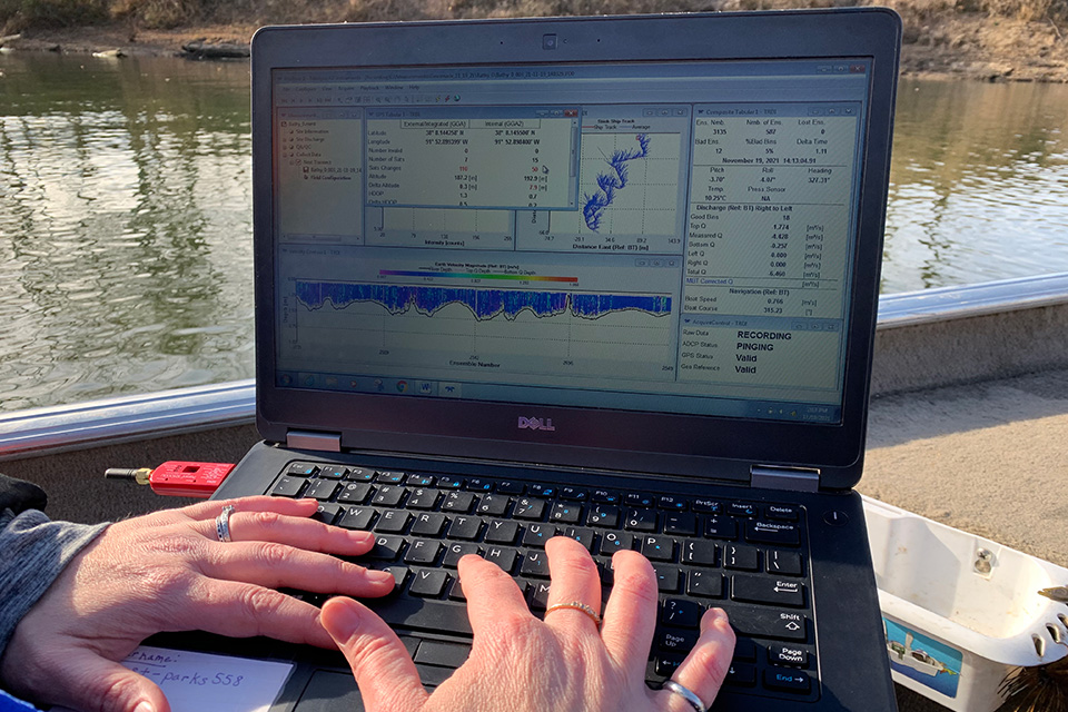 A computer screen shows the data collected at one of the bridge scour sites. A pair of hands type on a laptop keyboard, while the screen shows charts, graphs and data sets, situated with a river in the background.