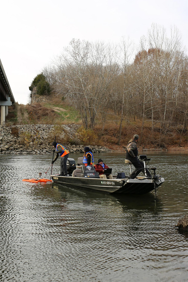 The SLU WATER Institute team adjusts equipment in the water at a study site. Four people are on a boat in a river, working with various pieces of equipment. Behind them, a shoreline with rocks and trees without leaves are visible.