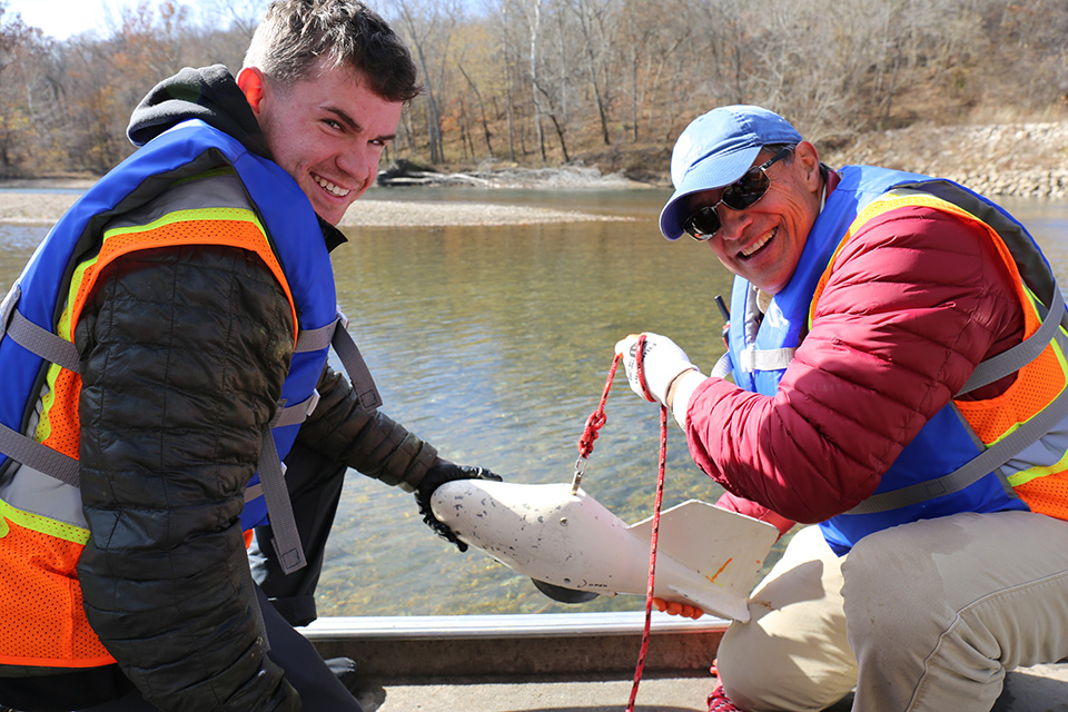 Luna (left) and Kickham (right) pose for a photo with some of the equipment used at a bridge scour study site. Two men wearing life jackets and reflective gear sit in a small boat on a river. They are holding a small piece of equipment on a rope. Rocky shore line with bare trees is visible in the background.