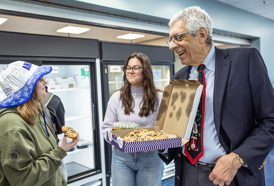 Pestello passes out cookies to students. Pestello holds a box of cookies and offers one to a female student wearing a SLU bucket hat while a woman in glasses looks on.